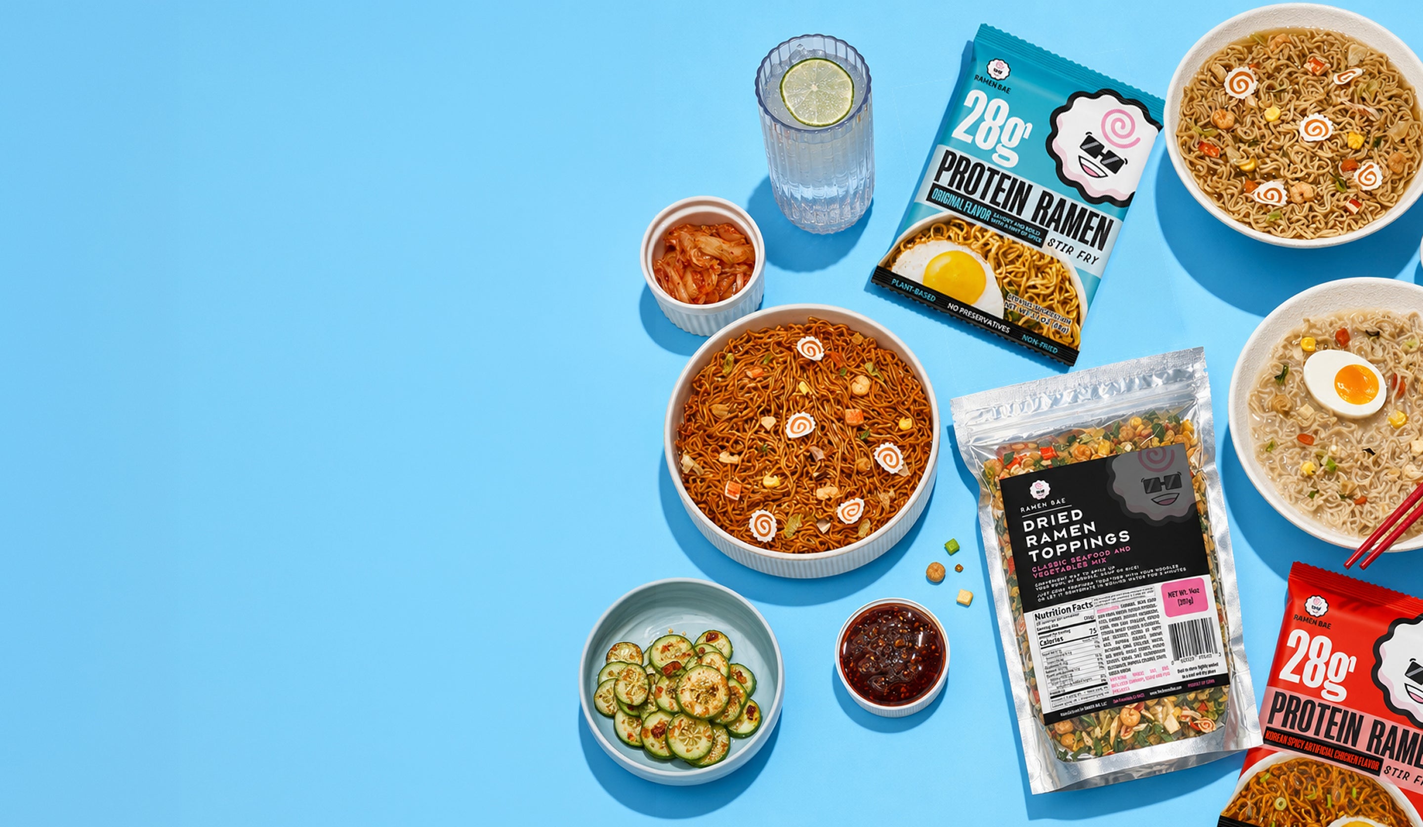 An overhead flat-lay of food products on a bright blue background, including bowls of ramen noodles, packaged dried ramen toppings, a protein ramen pouch, pickles, a drink with lime, and small condiment bowls.